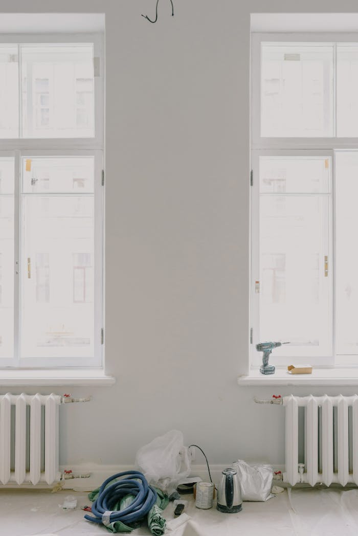 Interior of light room with white walls and new wooden windows above radiators during renovation with repairing tools and teapot located on floor